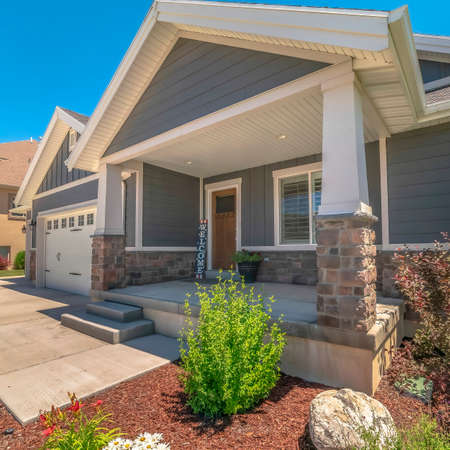 Square Home facade with view of yard and gable roofs over garage and front porch. A welcome sign is leaning against the wooden door and wall with gray siding and stone bricks.