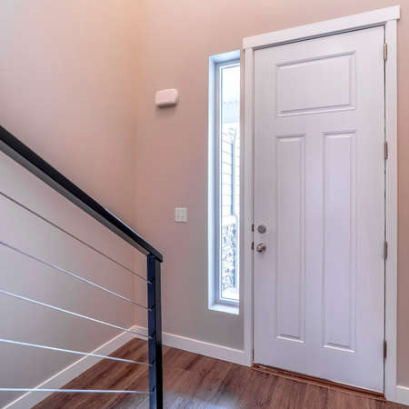 Square White hinged wooden front door and sidelight viewed from inside of home. Stairs handrail and...