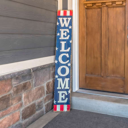Square frame Brown front door with glass pane and Welcome sign against gray and stone wall. Gray doormat, potted plant with flowers, and window can also seen at the house entrance.の写真素材