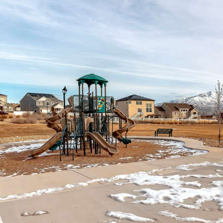Square frame Basketball court and playground at a park with melting snow on the gorund. The recreational area has a scenic view of houses, snowy mountain, and overcast sky.の写真素材