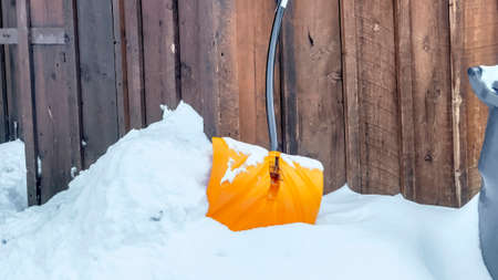 Panorama crop Yelllow snow shovel against snowed in ground and brown wooden wall of building. Spiked...