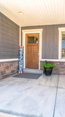 Vertical Home entrance with concrete porch brown wooden front door welcome sign. Window and potted plant with flowers can be seen against the wall with gray siding and stone bricks.の写真素材