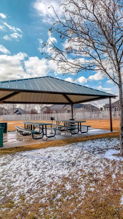 Vertical frame Pavilion with tables benches and melted snow against homes and cloudy blue sky. Trees that are starting to grow leaves can also be seen in this park.の写真素材
