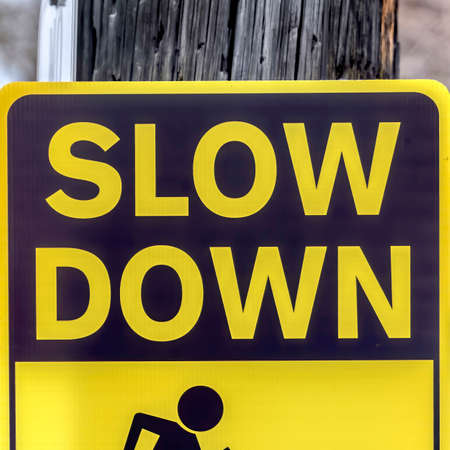Square frame Black and yellow Slow Down Kids And Pets At Play sign against gray wooden pole. Snowy landscape with trees and buildings can be seen in teh blurry background.の写真素材