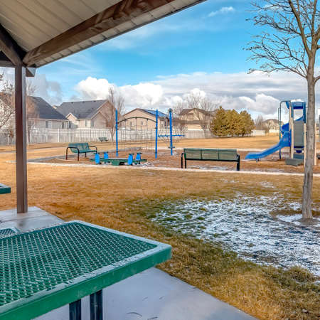 Square crop Green metal tables and benches inside a pavilion with melted snow on a sunny day. Playground, basketball courts, and houses can also be seen from the picnic area.の写真素材