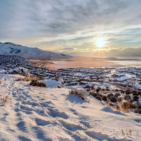 Square Snowy hill with scenic winter view of homes and lake and mountain in Draper Utah. Footprints on snow and bright sun against cloudy sky can also be seen in this beautiful landscape.の写真素材