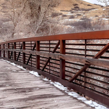 Square Bridge overlooking snowy hill landscape and trees against cloudy sky in winter. Melting snow can be seen on the wooden landing with metal railings on the sides.の写真素材