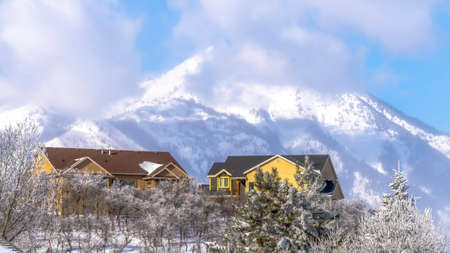 Panorama crop Distant snowy Wasatch Mountains with homes in foreground on a sunny winter day. Fristed trees, blue sky, and puffy clouds can also be seen in this picturesque scenery.の写真素材