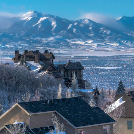 Square frame Snow falling on homes with sweeping view of valley and towering Wasatch Mountain. Fantastic winter landscape with mountain houses beneath blue sky.の写真素材