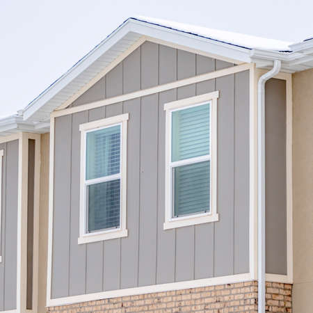 Square crop Snowy roof brick wall and vertical siding at exterior of townhome against sky. Residential landscape with view of the upper storey of townhouses.の写真素材