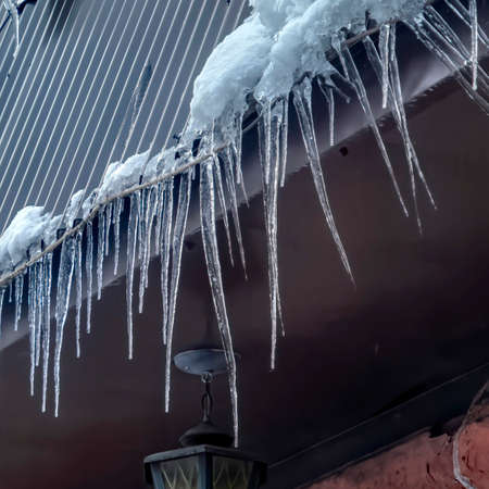 Square frame Spiked icicles at the edge of pitched gray roof with clumps of snow in winter. Frozen dripping water against ceiling lamp of house with stone wall.の写真素材