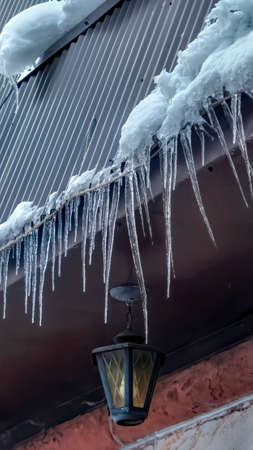 Vertical frame Spiked icicles at the edge of pitched gray roof with clumps of snow in winter. Frozen dripping water against ceiling lamp of house with stone wall.の写真素材