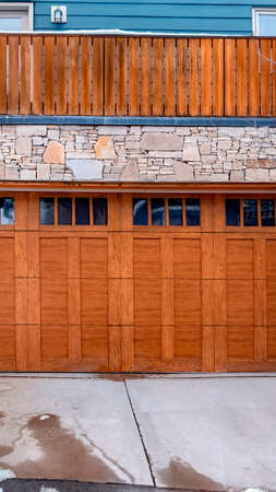 Vertical crop Brown wooden glass paned garage door against stone wall under balcony of home. Mounds of snow and and leafless tress can be seen around the driveway.の写真素材