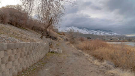 Panorama Dirt road along the retaining wall of slope overlooking lake and snowy mountain. Overcast sky can be seen over the nature and residential landscape.の写真素材