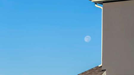 Panorama Close up of house exterior with blurry moon and blue sky in the background. The house has gray roof with white downspout at the edge of the wall.の写真素材