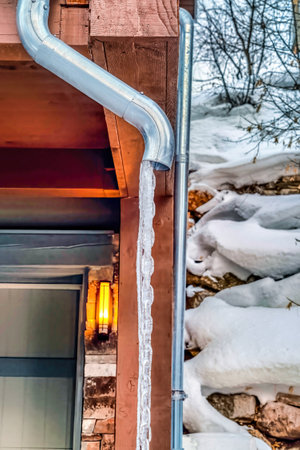 Roof drain pipe of home with frozen water against snowy and rocky hill slope
