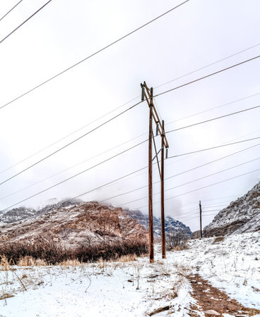 Dirt trail and power lines on snow covered terrain against hills and cloudy skyの写真素材