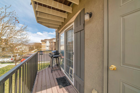 Barbecue grill on small balcony with roof overhang and huge window with shutters. The deck has a view of the neighborhood under blue sky on a sunny day.の写真素材