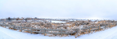 Snowy view of houses with grasses and trees with wooden fence