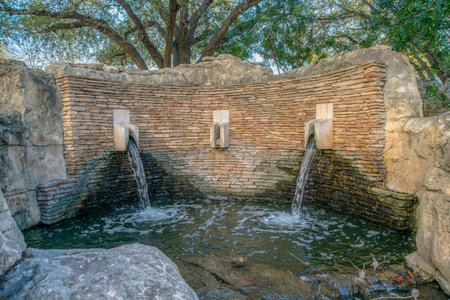 Water fountain with stone brick wall against trees in San Antonio Texas. Water from holes on the wall falls to the small pool with huge and ragged rocks.の写真素材