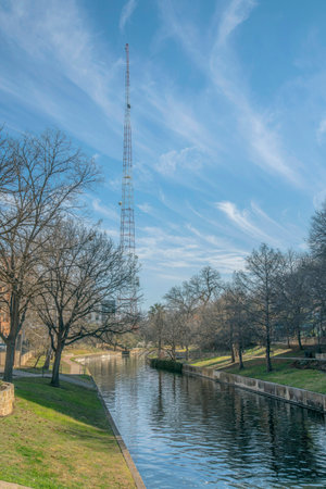 View of a tall signal tower near the River Walk at San Antonio River in Texas. There is a view of a grassy shore with leafless trees and a view of a tower against the cloudy sky background.の写真素材