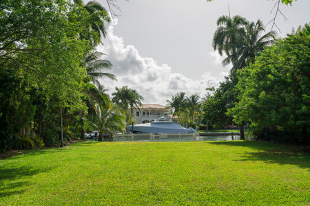 View of a yacht with cover from a yard with green lawn and fence at the bay in Miami, Florida. Views from a lawn with trees on the side across a large mansion with parked yacht at the front.の写真素材