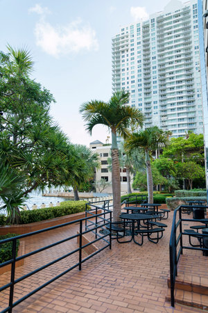 Outdoor lounge on deck near walkway with bricks metal barriers and tables with chairs at Miami, FL. There is a walkway with plants and trees near the water and view of building at the back.の写真素材