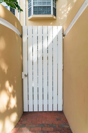 White door gate in the middle of building walls at Miami, Florida. Narrow entrance of a building with red bricks path in between the painted dark yellow walls with white trims.の写真素材