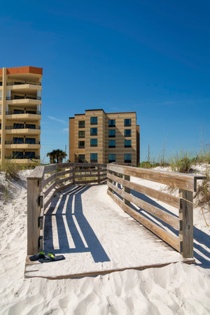 Sand covered wood pathway with handrails near the buildings against the clear blue sky at Destin, FL. There is a pair of slippers on the left near the wood railings on the left.の写真素材