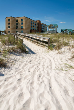 Path on sand heading to a boardwalk in the middle of sand dunes at the front of hotel- Destin, FL. Wood pathway in between the grasses on the white sand dunes and view of a hotel at the background.の写真素材