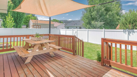 Panorama Whispy white clouds Wooden deck with a sunshade over the table with bench seats. Deck at the backyard of a house with a view of a lawn and white vinyl fence against the trees and neighborhood.の写真素材