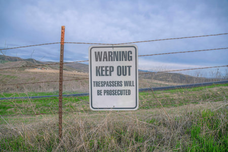 Hanging sign with Warning Keep Out Trespassers Will Be Prosecuted on a trail- San Clemente, CA. Hiking trail with warning sign hanging on a wire barrier against the mountain grassland and sky.の写真素材