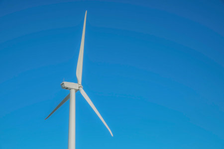 Close up view of a white windmill with three sharp blades against blue sky. The towering innovative windpump is an alternative source of green renewable energy.の写真素材