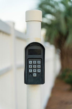 Destin, Florida- Digital keypad for automatic gate. Wireless automatic gate operator on a white pole outdoors with a blue background of a white fence and trees.の写真素材