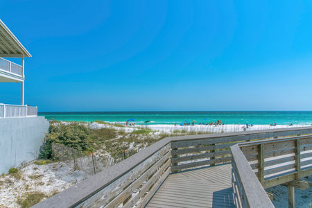 View of the beach with tourists on the shore from a boardwalk at Destin, Florida. There is a view of a beach house on the left with balconies near the tall grasses at the side and a waterfront view.の写真素材