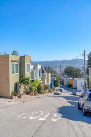 Suburban residential area on a sloped land in San Francisco, California. There is a concrete street in the middle with painted stop sign and parked vehicles and a view of the mountain at the back.の写真素材
