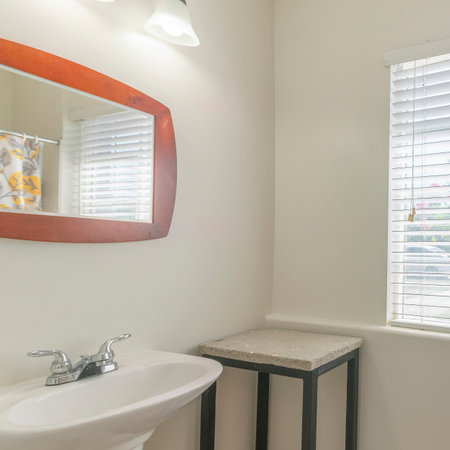Square Bathroom interior with table and pedestal sink with framed mirror. Small bathroom with printed shower curtain on the left beside the toilet near the window with blinds.の写真素材