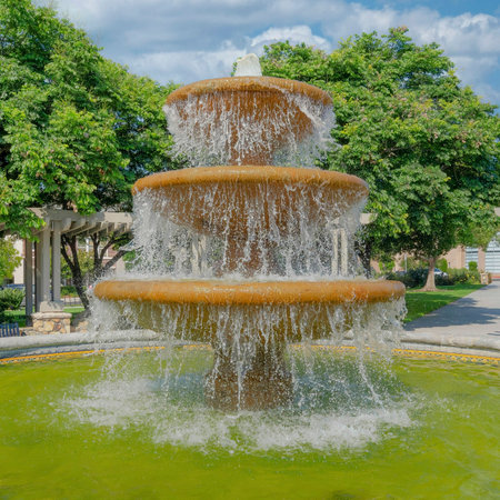 Square White puffy clouds Water fountain in the middle of a community plaza at San Marcos, California. There are green lawn and trees at the back of the walkway with pergola roof and a view of a building on the left.の写真素材