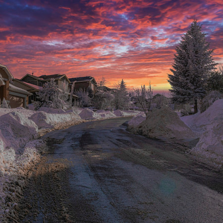 Square Dramatic sunset with clouds Uphill curved road cleared of snow in a residential area at Utah. There are thick snow piled up on the side of the road at the front of large houses with pine trees and garage.の写真素材