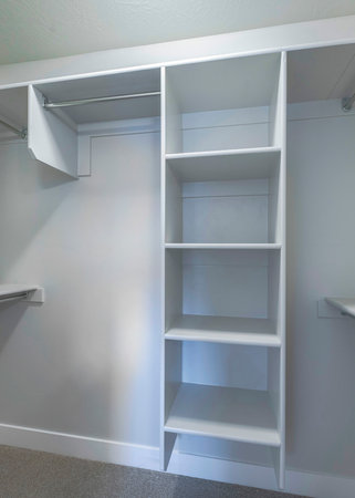 Vertical Empty small windowless walk in closet with open wardrobe. There are metal rods unter the wall mounted white wooden shelves and a light gray carpeted flooring.の写真素材