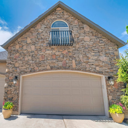 Square White puffy clouds Garage exterior with two garage doors and arched window on top. Three-car garage exterior with stone veneer siding and concrete driveway with two potted plants on the side.の写真素材