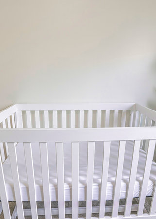 Vertical White wooden crib with white mattress in a white carpeted room. Crib with a linen basket on the right and potted small tree on the left.の写真素材