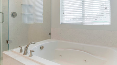 Panorama Bathtub and frameless shower stall with white marble wall surround. Interior of a bathroom with window and blinds near the bathtub with deck mounted faucet beside the stall with glass and shower head.の写真素材