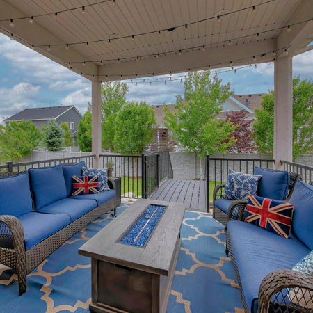 Square White puffy clouds Cozy outdoor deck with fire pit table and woven sofas with blue cushions. There is a blue carpet under the furnitures and an outdoor string lights on the ceiling, and a view of the backyard.の写真素材