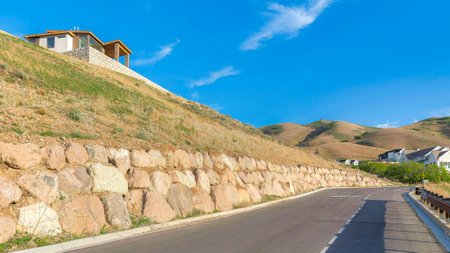 Panorama Paved road with roadside barrier beside the retaining wall of a man-made slope in Utah valley. There is a fenced house on top of the slope on the right and houses against the hills and clear sky.の写真素材