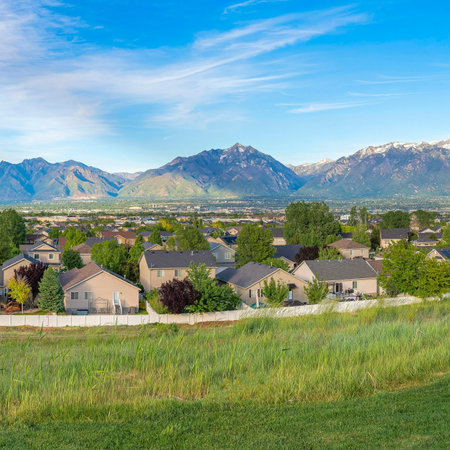 Square Whispy white clouds Panoramic view of a residential area with a mountain range background at Daybreak, Utah. There is a large field of grass at the front near the fenced residential area with trees outdoors.の写真素材
