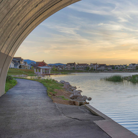 Square Whispy white clouds Under an arched bridge over Oquirrh Lake at Daybreak, Utah. There are two paths beside the lake with buoyancy balls and a view of residential houses against the sunset horizon sky at the back.の写真素材