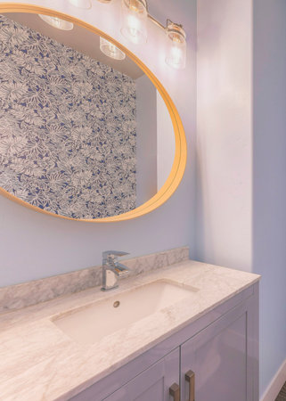 Vertical Powder blue bathroom interior with floral wall paper. There is a toilet with portrait picture frame at the back and a vanity sink with marble counter and framed oval mirror.の写真素材