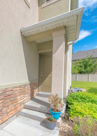 Vertical Whispy white clouds Entrance of a house at the side of the wall with stone veneer. Front door with potted plants on the concrete steps beside the bushes and green lawn against the vinyl fence and neighbor's house.の写真素材