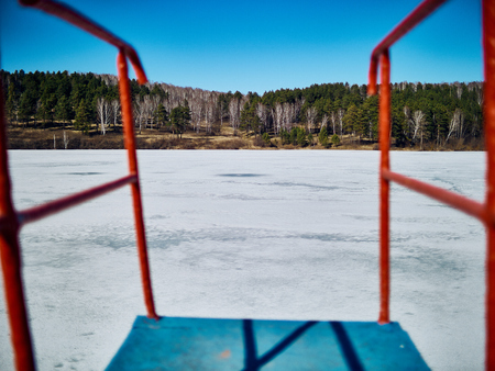 Cold Siberian landscape of a lake strained in the ice. View of the road on the back side of the frozen lake through the handrails of the tower for diving.の写真素材